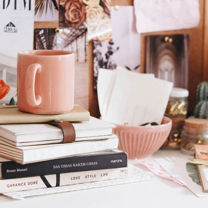 A pink mug sits on a stack of books and notebooks on a cluttered desk with various decor items and photos in the background.