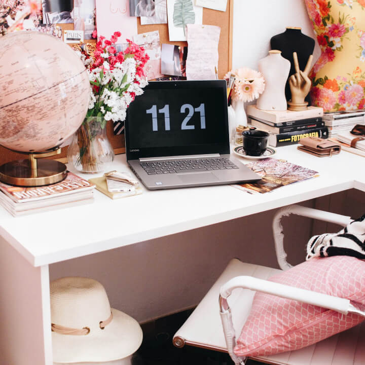 A cluttered desk with a laptop displaying "11:21," a globe, flowers, books, a coffee cup, and a hat.