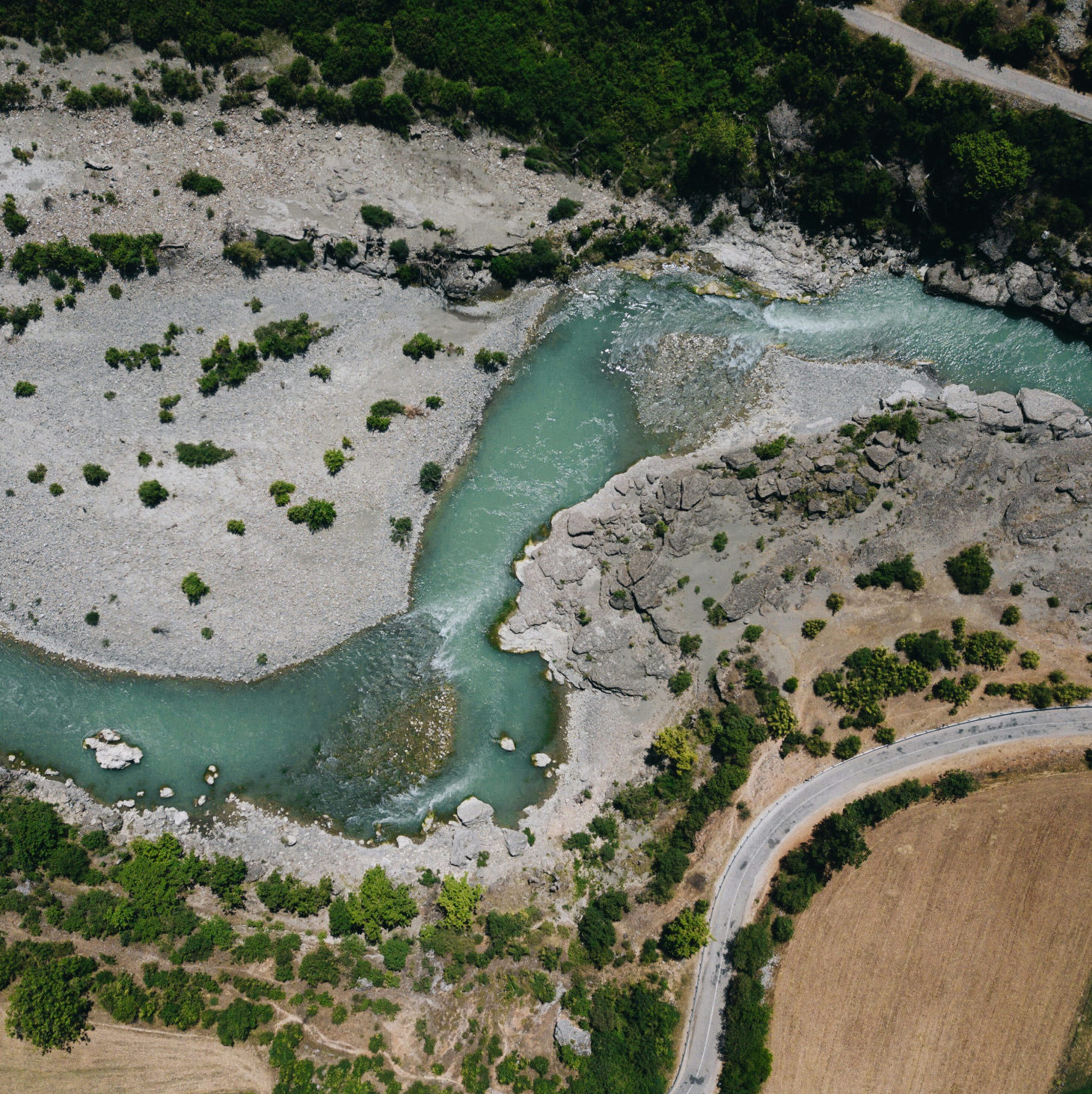 Aerial view of a winding turquoise river flowing through rocky terrain and surrounded by greenery, with a road curving along a dry field.